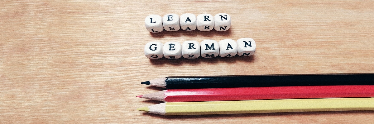 Des dés avec des lettres "Apprendre l'allemand" à côté de 3 crayons en noir, rouge et jaune