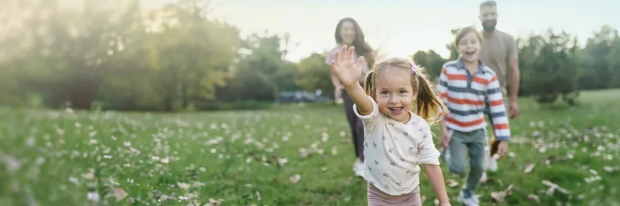 Child running and waving in a sunny field, family following behind.