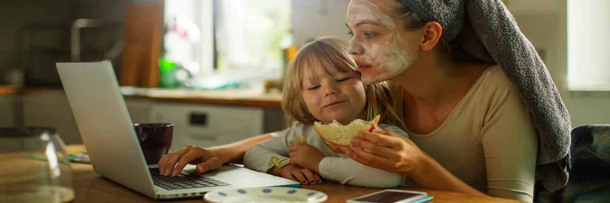A multitasking parent sits in front of a laptop, child in her lap, sandwich in hand, towel in hair