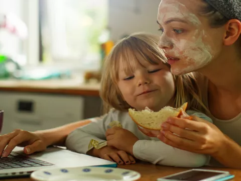 A multitasking parent sits in front of a laptop, child in her lap, sandwich in hand, towel in hair