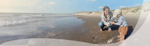 Parent and child playing on a sunny beach, waves in the background.