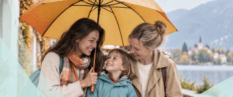 Family under an umbrella, smiling by a lake with mountains in the background.