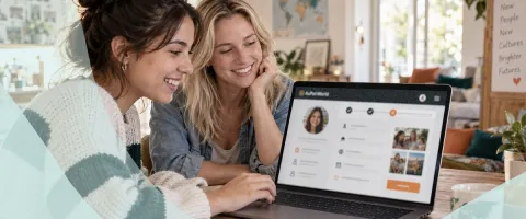 Two women smiling, looking at a laptop screen in a cozy room.