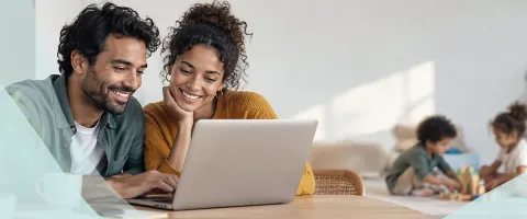 Couple smiling at a laptop with children playing in the background.