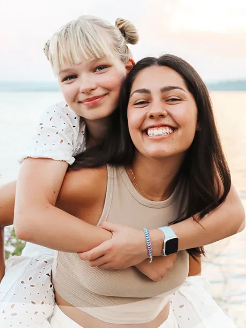 Two smiling women, one giving a piggyback ride by a lake.