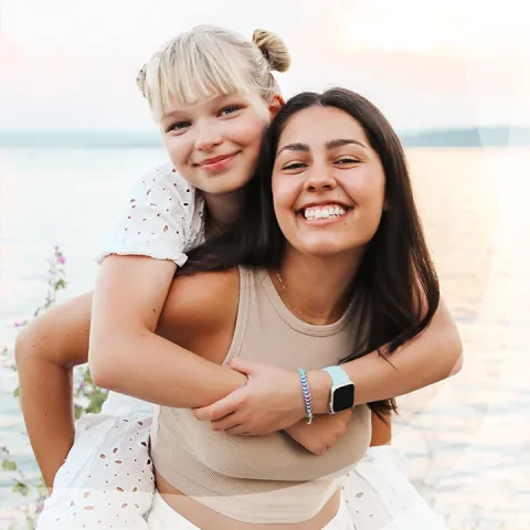 Two smiling women, one giving a piggyback ride by a lake.